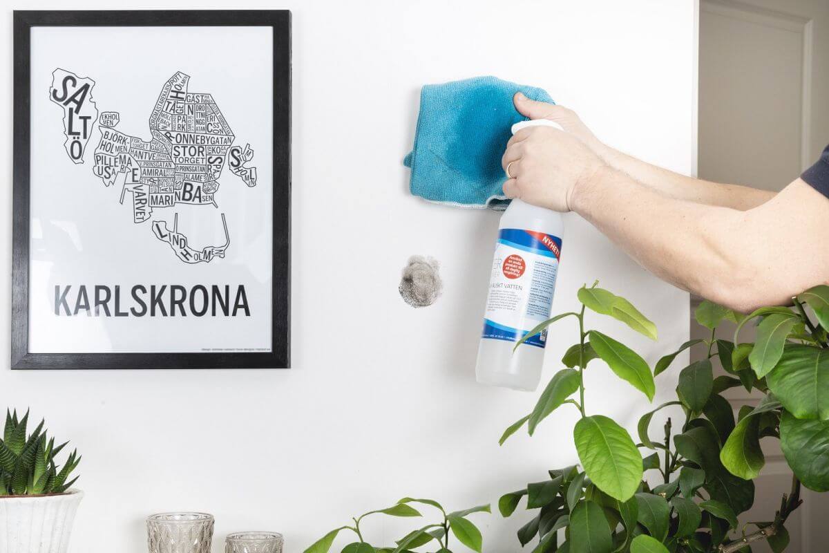 Ręka trzymająca niebieską szmatkę i butelkę ze środkiem czyszczącym, ściana z zabrudzeniem i ramka z mapą miasta Karlskrona oraz kilka roślin doniczkowych na dole.