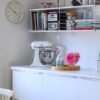 Biała kuchnia ze ścianą z półkami z książkami i kuchennymi sprzętami, stołem z mikserem, tacą z kwiatem i spryskiwaczem oraz doniczką z kaktusem, a nad blatem zegar ścienny.