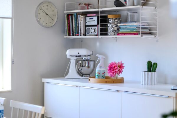 Biała kuchnia ze ścianą z półkami z książkami i kuchennymi sprzętami, stołem z mikserem, tacą z kwiatem i spryskiwaczem oraz doniczką z kaktusem, a nad blatem zegar ścienny.