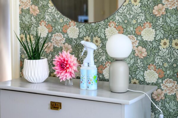 Biała komoda z lustrem na ścianie, na której stoją doniczka z zieloną rośliną, sztuczny różowy kwiat, dwie butelki z rozpylaczami i nowoczesna lampa z białą kulistą kloszem na beżowej podstawie, w tle tapeta w zielono-brązowe kwiaty.