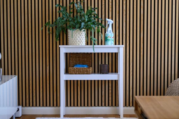Biała konsola z rośliną doniczkową, sprayem i wiklinowym koszykiem na tle ściany z pionowych drewnianych listew