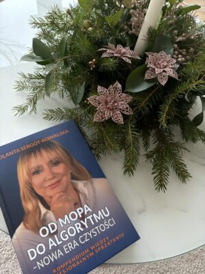 Książka "Od mopa do algorytmu - Nowa era czystości" obok świątecznej dekoracji z zielonymi gałązkami, różowymi ozdobnymi kwiatami i białą świecą na szklanym stole.